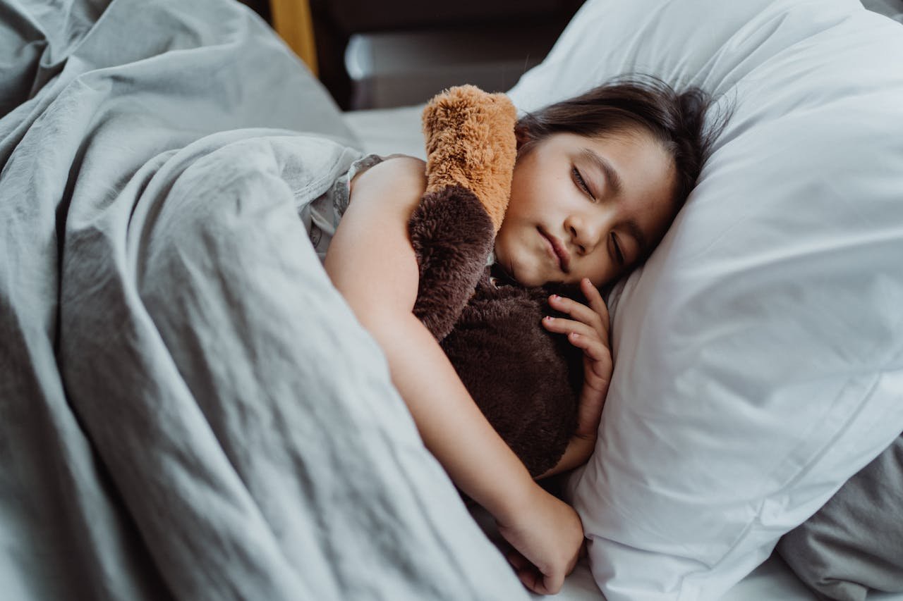 portfolio-06 A young girl peacefully sleeping in bed with a stuffed teddy bear, showcasing tranquility and comfort.