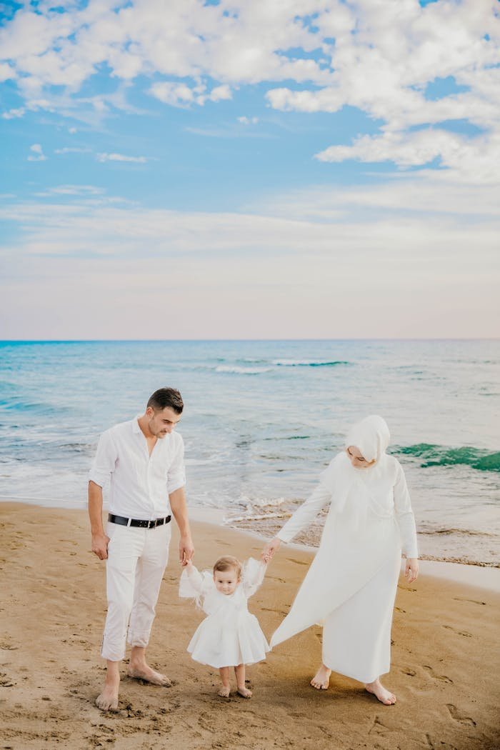 core-values Full length of Muslim couple in stylish white clothes holding hands of little daughter while walking on sandy coast near wavy ocean in sunny day